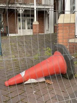 Orange plastic cone lying on the ground behind metal construction fence Stock Photos