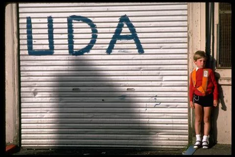 Orange-sashed boy standing next to store gate w. UDA graffiti, during Orange Par Stock Photos