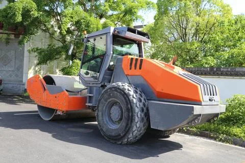 Orange single-drum road roller parked on asphalt beneath leafy trees in bri.. Stock Photos