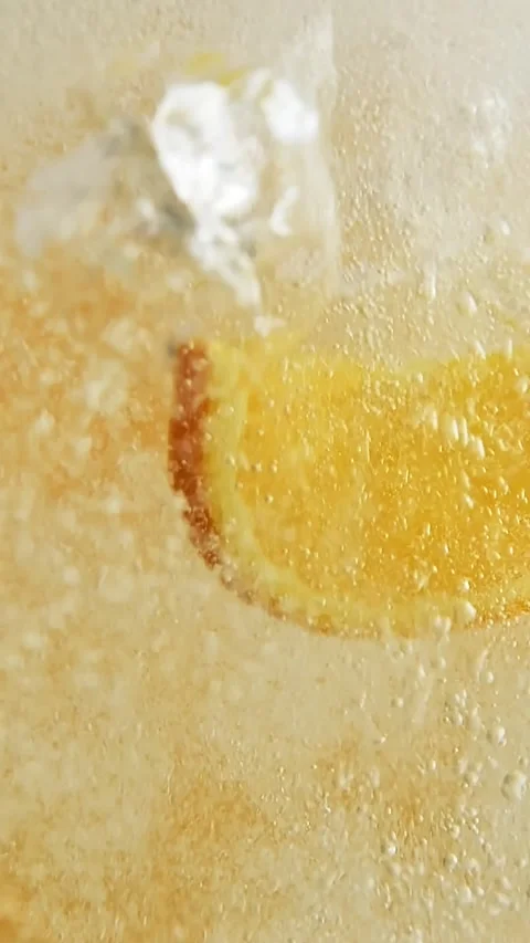Orange slice drops into fizzy golden drink with ice cubes, creating sparkling Stock Footage 306283053