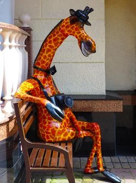 Orange smiling giraffe statue with a camera around its neck sits on a bench Fotos Stock