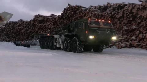 Oshkosh low-loader vehicle driving on snow past stockpile of logs Video stock 265154517