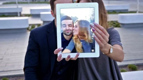 Ouple  sitting on a bench, trying to take a  selfie. Girl is kissing his boy Stock Footage 55390782