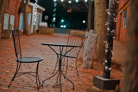 Outdoor Cafe With Porch Lights Over A Chair & Table At Night On A Brick Pathway Stock Photos