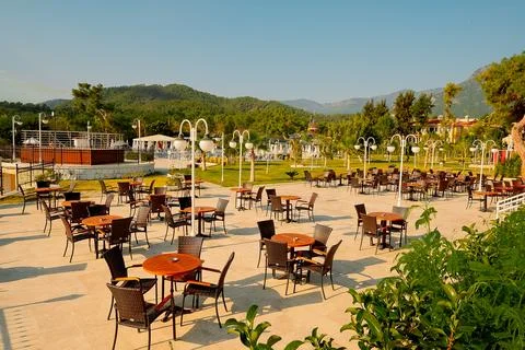 An outdoor dining area with empty tables and chairs. Sunlight shines on green Stock Photos