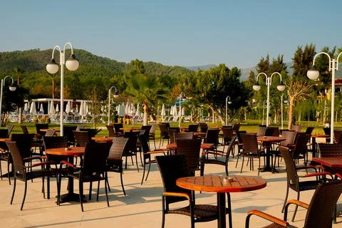 An outdoor dining area with empty tables and chairs. Sunlight shines on green Stock Photos
