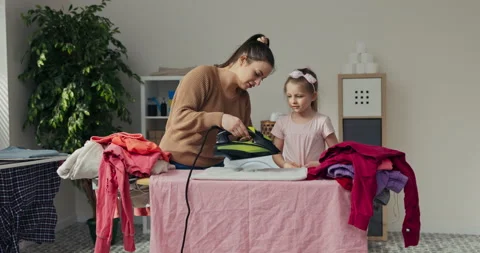 Outside the window is sunny day. Young mother shows daughter how to iron clothes Stock Footage 218316953
