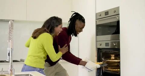 Oven timer ringing, Diverse couple reaching into oven in kitchen pulling Stock Footage 317661705