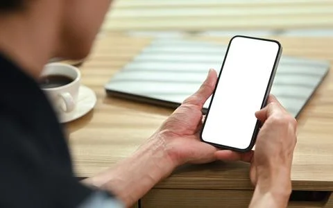 Over shoulder view of man using smart phone on wooden desk. Blank screen for Stock Photos