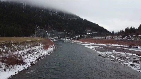 Over a wide flowing river between snow covered rocky shore with tree covered Stock Footage 125054192