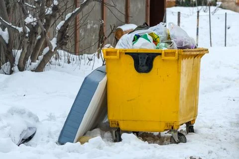 Overflowing yellow garbage container with trash bags in snowy winter yard Stock Photos
