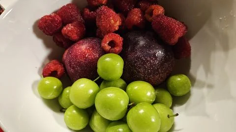 Overhead Close-Up of Raspberries, Plums and Green Cherries in a White Bowl Stock Photos