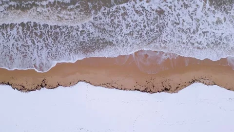 An overhead perspective of a winter shoreline where foamy ocean waves meet snowy Stock Footage 302445054