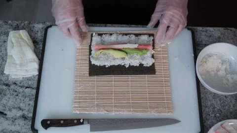 OVERHEAD VIEW OF GLOVED HANDS ADDING FILLINGS AND ROLLING SUSHI ON BAMBOO MAT Stock Footage 336947321