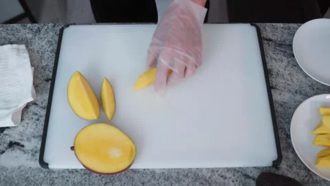 OVERHEAD VIEW OF GLOVED HANDS SLICING MANGO ON CUTTING BOARD FOR FOOD PREP 스톡 동영상 336947566