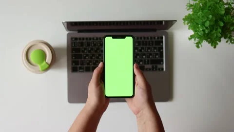 Overhead view of hands holding a green screen smartphone near a laptop and plant Stock Footage 337173313
