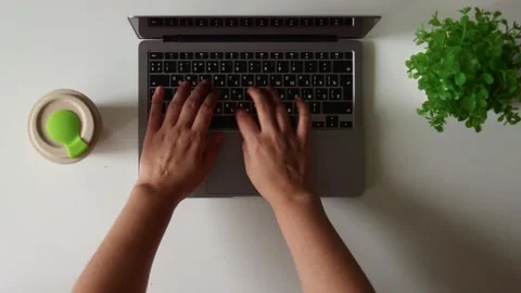 Overhead view of hands typing on a laptop keyboard with a plant and cup nearby Stock Footage 337173546