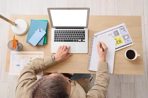 Overhead view of man working on laptop and taking notes at wooden desk with c Stock Photos