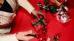 Overhead view of a woman using scissors, cutting green ribbon, making curly  ornaments while decorating a Christmas gift, wrapped in red wrapping paper.  Christmas time. Happy New Year. Diy present Stock Footage