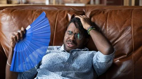 Overheated afro american man recline on couch use hand fan Stock Photos