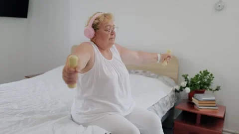 Overweight elderly lady doing dumbbell raises sitting on bed at home fitness at 库存影片 337909279