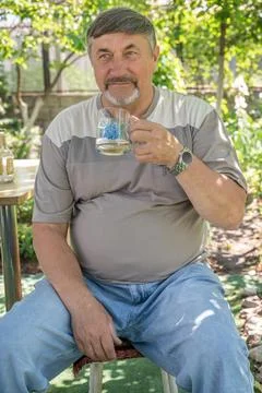 An overweight elderly man drinking while sitting on a chair in the garden. .. Stock Photos