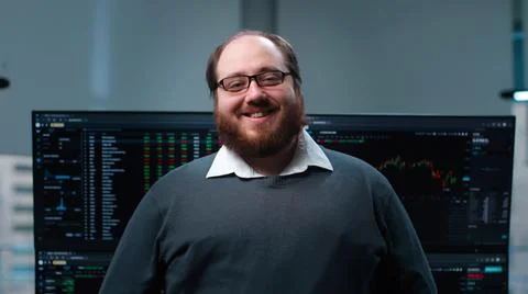 Overweight it-programmer smiling at camera with multiple screens with charts on Foto stock