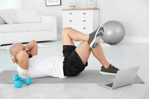 Overweight man doing exercise while watching tutorial on laptop at home Stock Photos