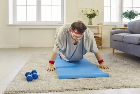 Overweight man doing push-ups on yoga mat at home, humorous fitness motivation Stock Photos