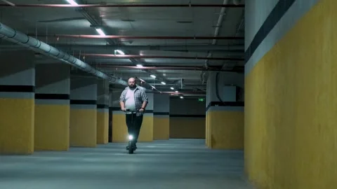 Overweight man riding e-scooter in underground parking. Male replace a car on Stock Footage 255833871