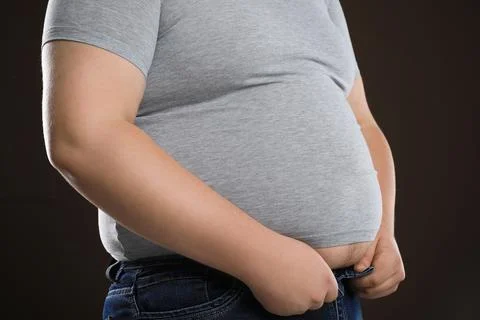 Overweight man trying to button up tight jeans on dark brown background, clos Stock Photos