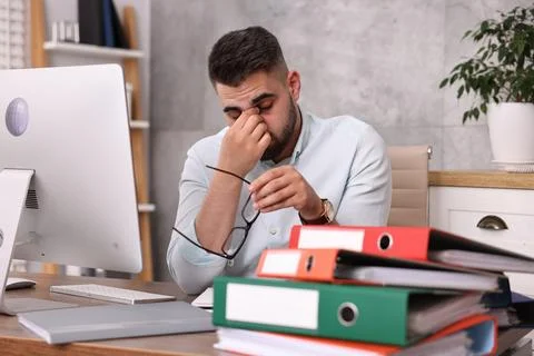 Overwhelmed man sitting at table with computer and stack of folders in office Stock Photos