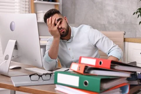 Overwhelmed man sitting at table with computer and stack of folders in office Stock Photos