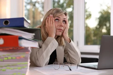 Overwhelmed woman sitting at table with stack of documents and folders in o.. Foto stock