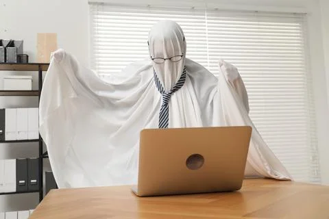 Overworked ghost. Man covered with white sheet using laptop at wooden table.. 스톡 사진
