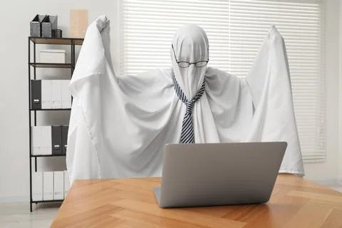 Overworked ghost. Man covered with white sheet using laptop at wooden table.. Stockfoto's
