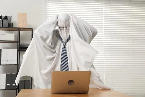 Overworked ghost. Man covered with white sheet using laptop at wooden table.. 写真素材