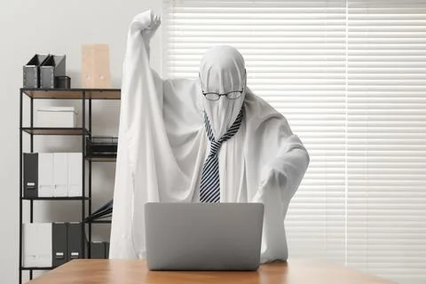 Overworked ghost. Man covered with white sheet using laptop at wooden table.. Stockfoto's