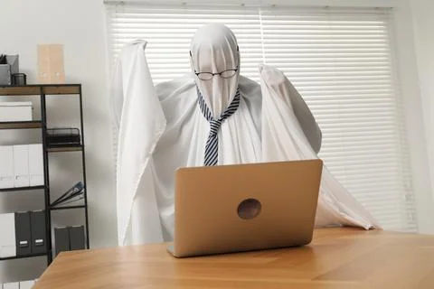 Overworked ghost. Man covered with white sheet using laptop at wooden table.. 스톡 사진