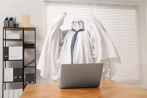 Overworked ghost. Man covered with white sheet using laptop at wooden table.. 写真素材