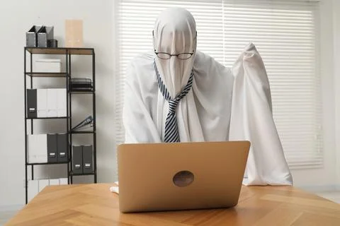 Overworked ghost. Man covered with white sheet using laptop at wooden table.. 스톡 사진