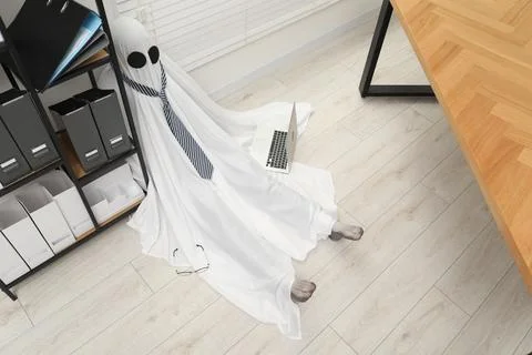 Overworked ghost. Man in white sheet with laptop on floor in office, above vi 스톡 사진