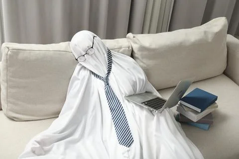 Overworked ghost. Man in white sheet with laptop and books on sofa at home 스톡 사진