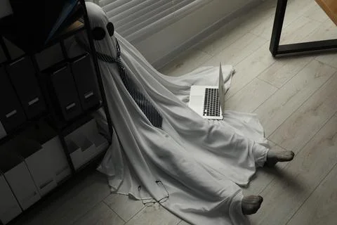 Overworked ghost. Man in white sheet with laptop on floor in office, above vi Stockfoto's