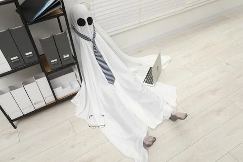 Overworked ghost. Man in white sheet with laptop on floor in office, above vi 스톡 사진