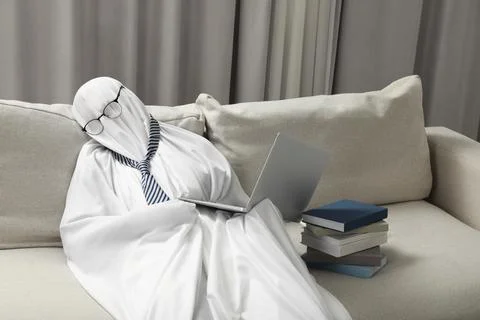 Overworked ghost. Man in white sheet with laptop and books on sofa at home 스톡 사진