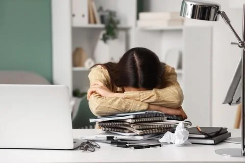 Overworked young woman sleeping on a stack of notebooks and papers at a messy Stock Photos