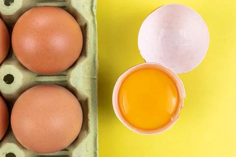 Pack of brown eggs with one broken open egg with raw yolk and shell on yellow Stock Photos