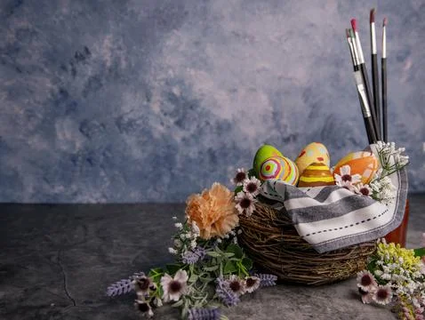 Paint brushes and colored Easter eggs in a straw corp with flowers Still life Stock Photos
