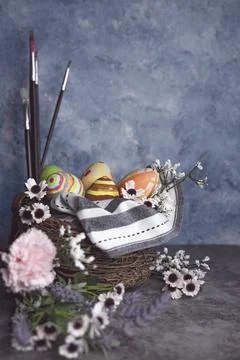 Paint brushes and colored Easter eggs in a straw corp with flowers Still life Stock Photos